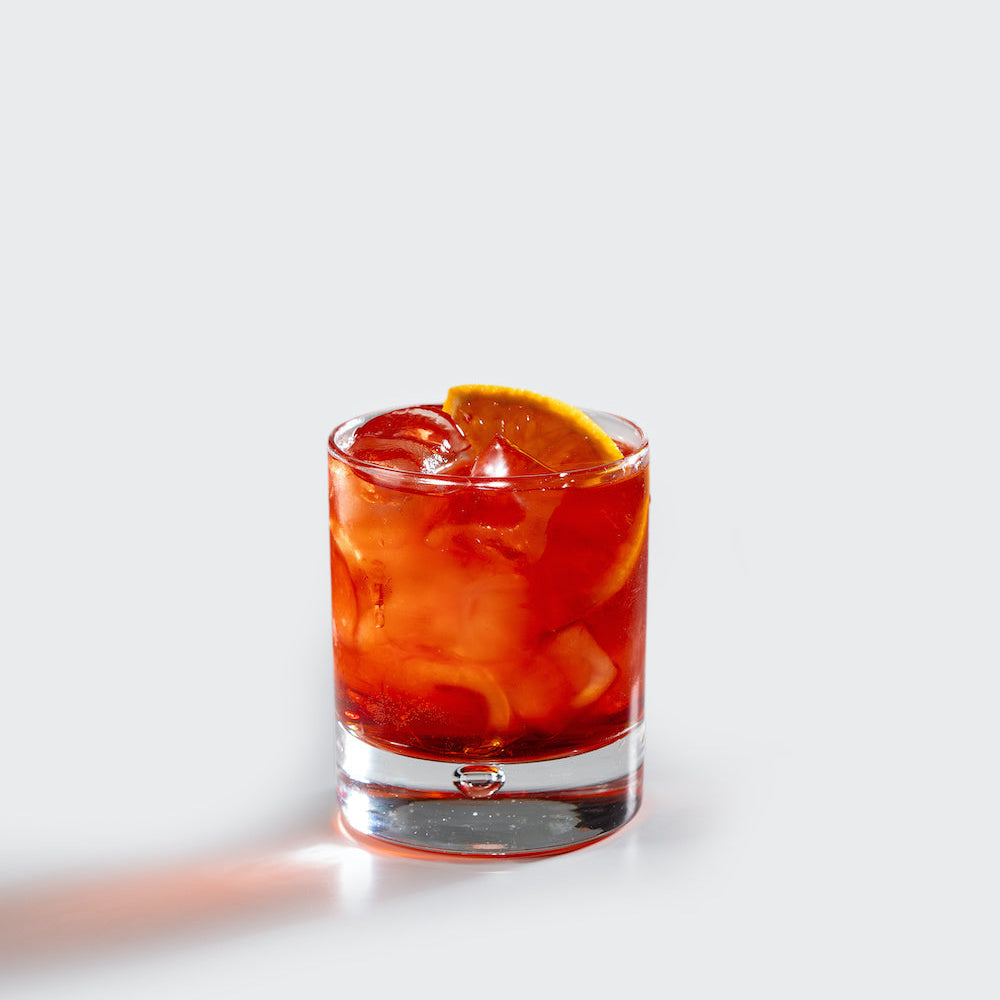 A Boulevardier cocktail served in a clear rocks glass over a large ice cube, garnished with a Boozy Cocktail Cherry® and orange peel