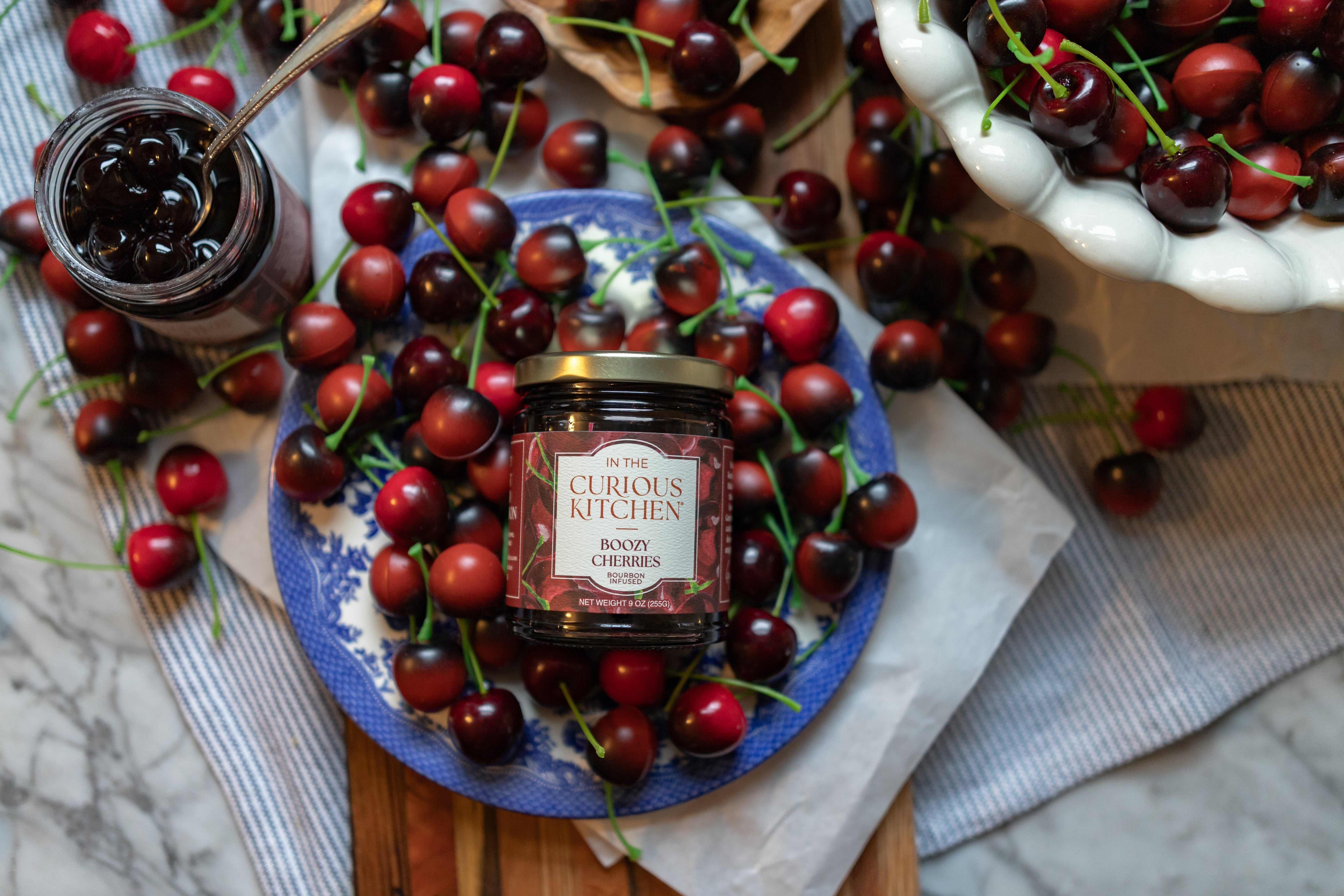 A jar of Boozy Cocktail Cherries® from In the Curious Kitchen resting on a bed of fresh dark cherries
