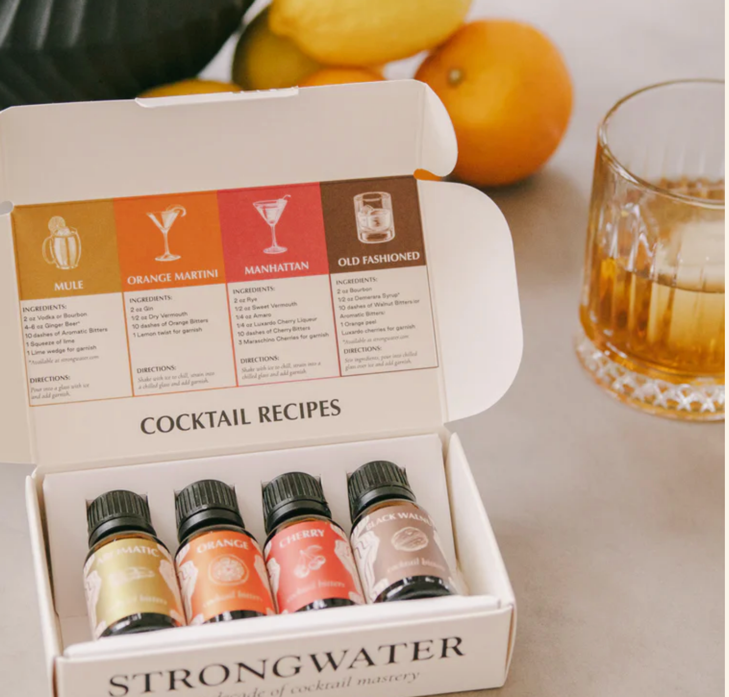 Box of Strongwater cocktail ingredients with cocktail recipes on a table with lemons and a glass.