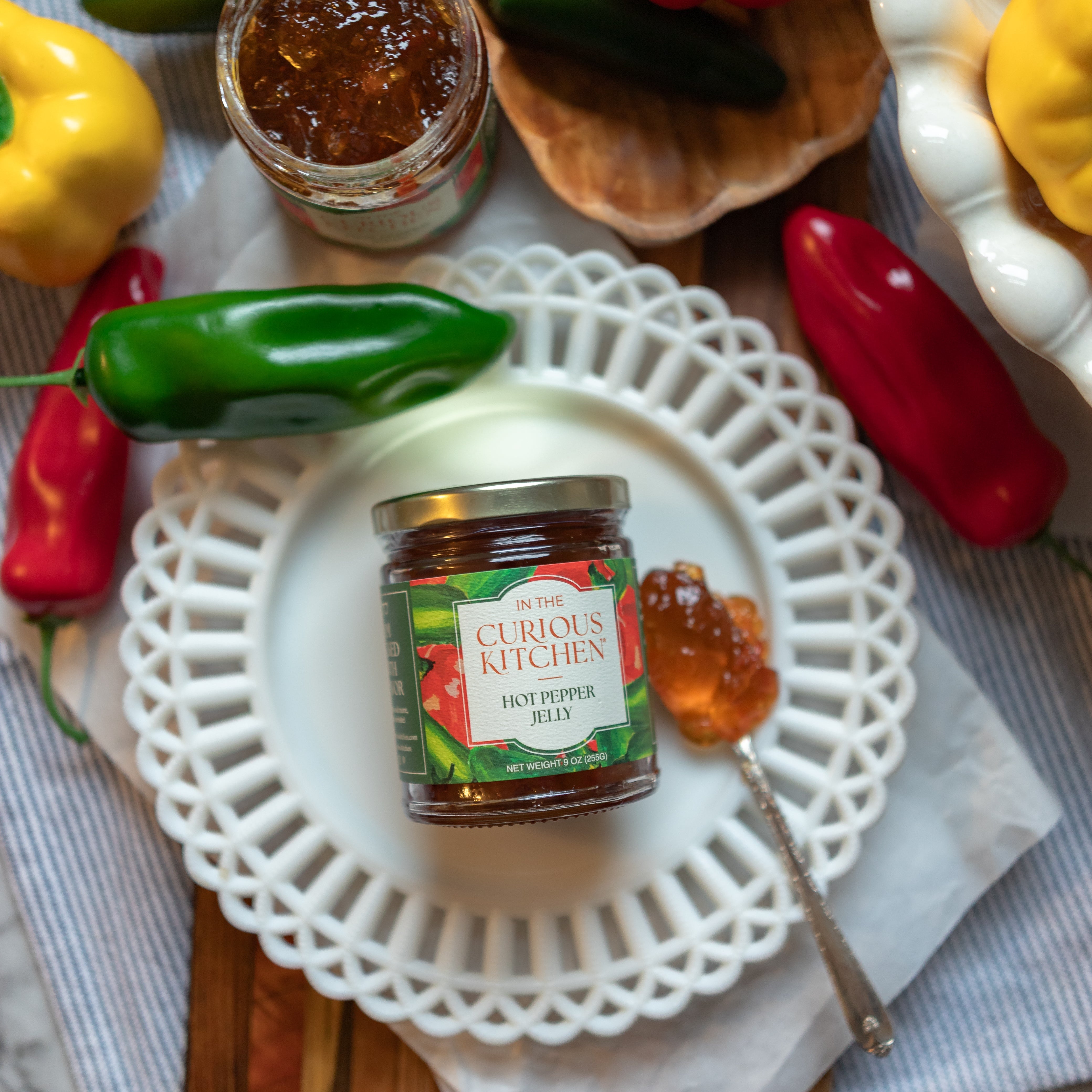Jar of Curious Kitchen hot pepper jelly on a white plate with peppers and honey in the background.