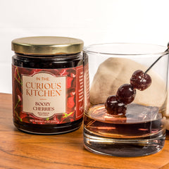 In the Curious Kitchen Boozy Cherries jar beside a bourbon Old Fashioned garnished with cocktail cherries on a pick