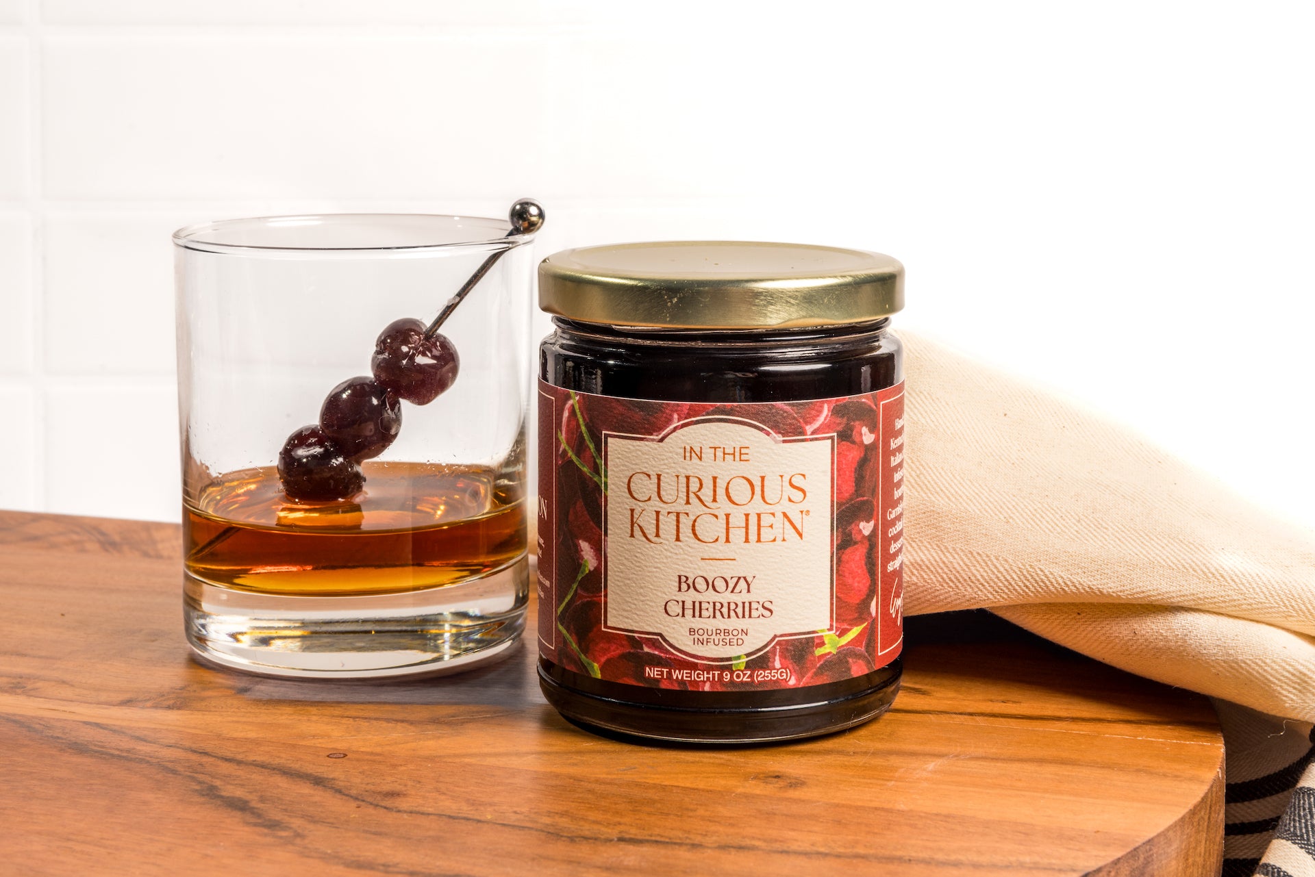 Jar of 'In the Curious Kitchen' boozy cherries with a glass of whiskey on a wooden surface.