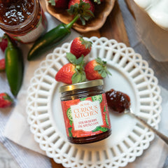 Jar of strawberry jalapeño jam from 'In the Curious Kitchen' on a decorative plate with strawberries and a cup of coffee.